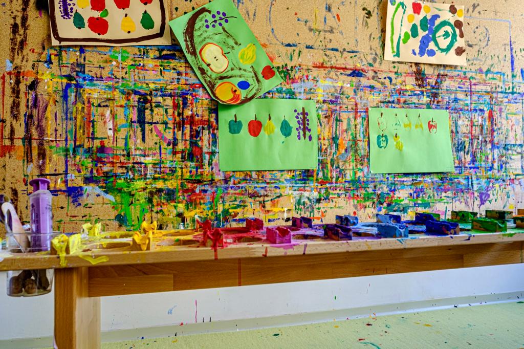 Maltisch in einer Kita, dahinter eine bunt bemalte Wand mit Bildern von Kindern