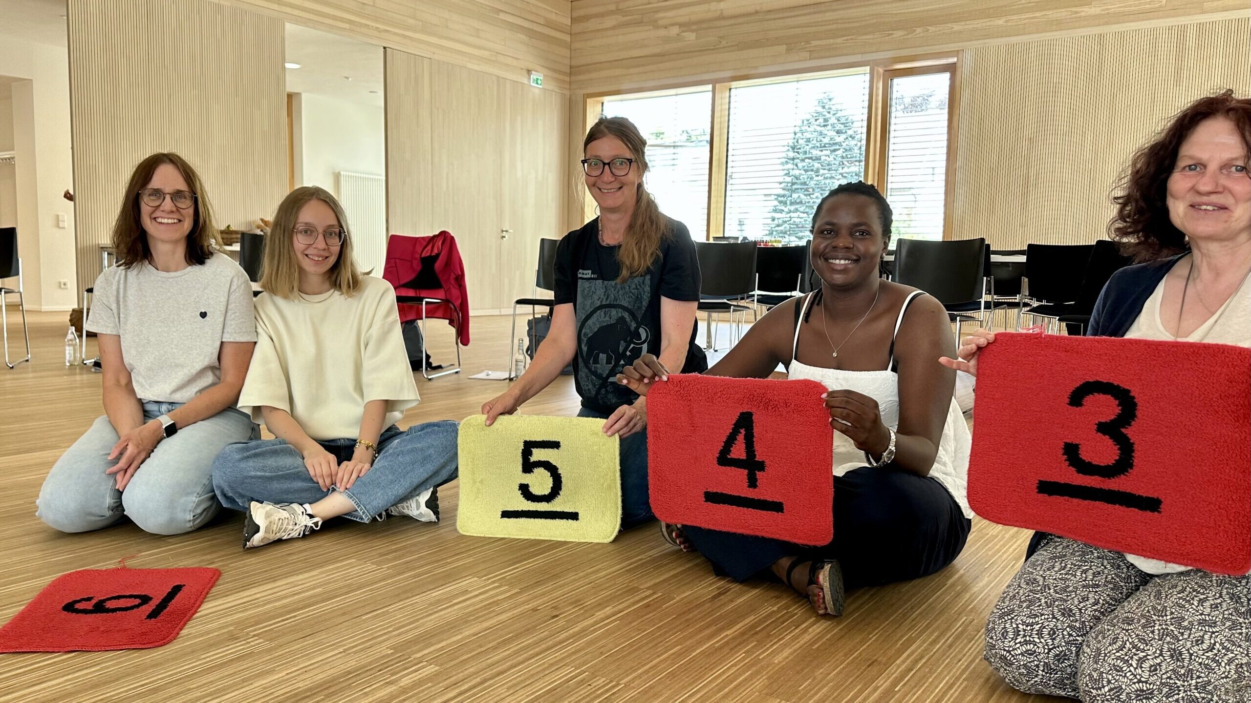 Fünf Teilnehmerinnen sitzen auf dem Boden und halten Matten mit den Zahlen 3, 4 und 5